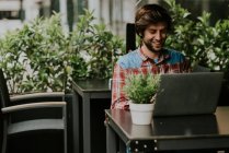 Portrait d'un homme barbu assis à la table de terrasse d'un café avec une plante en pot et utilisant un ordinateur portable — Photo de stock