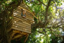 Bottom view of blonde boy looking out of wooden treehouse window on sunny day — Stock Photo