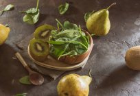 Plat en bois rempli de feuilles d'épinards frais servi avec kiwi tranché sur une assiette à table avec des poires et une cuillère en bois — Photo de stock