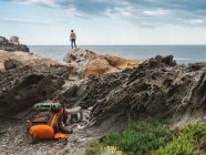 Mochila de viagem deitada em rochas sobre o viajante de pé em penhascos de rocha e admirando a paisagem oceânica — Fotografia de Stock