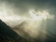 Paisagem pitoresca de montanhas enevoadas iluminadas com raios de sol lutando através de nuvens pesadas no céu sombrio . — Fotografia de Stock