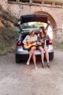 Girls sitting with guitar in car trunk — Stock Photo