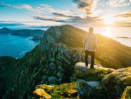Back view of man standing on rock on background of mountains and ocean in bright sunlight. — Stock Photo