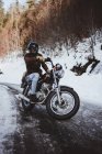 Man in helmet posing on chrome motorcycle with background of bare trees on snowy road. — Stock Photo
