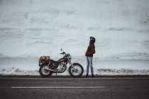 Hombre en chaqueta Alaska de pie junto a la motocicleta en carretera nevada - foto de stock