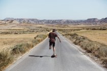 Visão traseira do homem em camiseta e shorts andando de skate na estrada da pradaria no dia ensolarado . — Fotografia de Stock