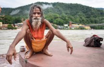 Obdachloser erwachsener indischer bärtiger Mann sitzt am Fluss und schaut in die Kamera. — Stockfoto
