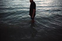 Crop man en camiseta negra y pantalones cortos de pie hasta las rodillas en el agua . - foto de stock