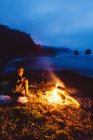 Uomo in pantaloncini seduto accanto al falò in riva al mare di notte e guardando fuoco fiammeggiante . — Foto stock