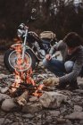Man keeping campfire burning on background of parked motorcycle on rocky shore. — Stock Photo