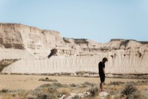 Vista laterale dell'uomo in maglietta nera e pantaloncini in piedi nel deserto nella giornata di sole e guardando a terra . — Foto stock