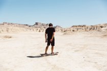 Vista trasera del hombre en camiseta negra y pantalones cortos de montar monopatín en el desierto en el día soleado . - foto de stock