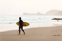 Visão traseira do surfista criança caminhando para ondas na costa arenosa à beira-mar . — Fotografia de Stock