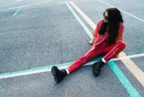 Brunette girl in red clothes sitting on asphalt at playground and looking away — Stock Photo