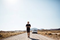 Homme barbu tenant la caméra et marchant le long de la route du désert loin de la voiture garée . — Photo de stock