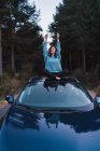 Усміхнена дівчина в синій сорочці сидить на вершині автомобіля з піднятими руками на лісовій дорозі — стокове фото