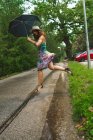 Vista lateral de la mujer morena saltando con paraguas sobre la paleta en el camino del parque . - foto de stock