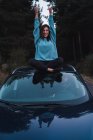 Mädchen in blauem Sweatshirt sitzt mit überkreuzten Beinen und erhobenen Armen auf Auto — Stockfoto