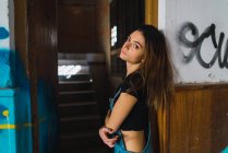 Vue latérale de la fille brune posant dans un bâtiment abandonné et regardant la caméra — Photo de stock