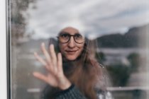 Retrato de mujer con gafas y sombrero blanco y cristal tocando detrás de la ventana - foto de stock