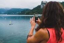 Visão traseira de mulher tomando tiros com smartphone de lago em montanhas . — Fotografia de Stock