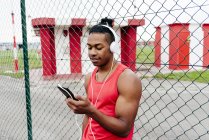 Portrait of sportsman in headphones leaning on fence and browsing smartphone — Stock Photo