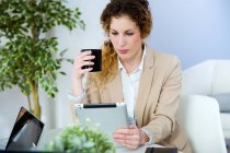 Retrato de mujer de negocios con taza de café usando tableta en la oficina moderna . - foto de stock