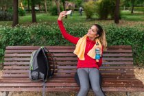 Молода усміхнена жінка з рушником на плечах приймає селфі зі смартфоном, сидячи на лавці — стокове фото