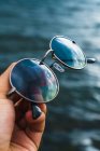 Crop hand holding sunglasses reflecting blue sky over sea waves — Stock Photo