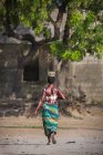 Вид сзади на племенную женщину, идущую с чашей на голове в деревне — стоковое фото