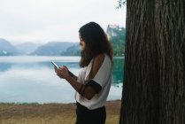 Vista laterale di bruna donna che naviga smartphone sulla riva del lago . — Foto stock