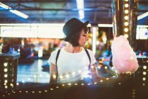 Vista lateral de la joven mujer sosteniendo hilo de azúcar rosa y posando románticamente en las luces del parque de atracciones . - foto de stock