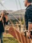 Woman with vintage camera taking shot of man with guitar case at countryside field — Stock Photo
