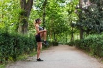 Вид збоку на спортивну дівчину, що розігріває ногу перед бігом на парковій алеї — стокове фото