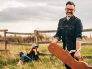 Woman with vintage camera leaning on rural fence and taking shot of smiling man posing with guitar case and looking at camera — Stock Photo
