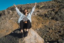 Low angle portrait of woman wearing denim coat sitting on stone at rocky slope with arms up — Stock Photo