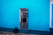 Kuba - 27. August 2016: Mann sitzt auf Gehweg und lehnt sich an blaue Wand mit dekorativer Metalltür. — Stockfoto