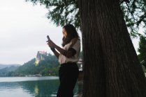 Вид сбоку на брюнетку, стоящую у дерева и просматривающую смартфон — стоковое фото
