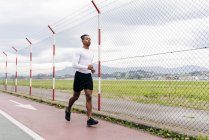 Hombre en ropa deportiva corriendo a lo largo de la cerca en el día nublado - foto de stock