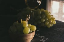 Vista de cerca del racimo de uvas verdes con pinchos en un tazón al lado de un vaso de vino blanco - foto de stock