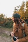 Portrait of brunette woman wearing cap and coat holding camera and looking aside at countryside — Stock Photo