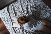Вид сверху на девушку в рубашке, читающую книгу на кровати — стоковое фото