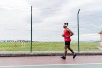 Vue latérale du sportif en casque jogging le long de la clôture — Photo de stock