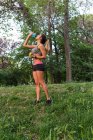 Спортивная девушка с яблоком в руке питьевой воды после тренировки на газоне парка — стоковое фото