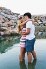 Vista lateral do casal abraçando em pé na água e beijando sobre o fundo de pedras costeiras — Fotografia de Stock