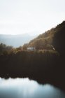 Vista lejana a la pequeña casa en la orilla del lago con montañas verdes en la niebla en el fondo  . - foto de stock