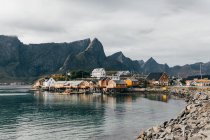 Small houses of mountain village standing on lake shore on background of peaks and cloudy sky. — Stock Photo