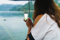 Vista trasera de mujer con paraguas tomando fotos con smartphone del lago en las montañas . - foto de stock