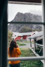 Вид ззаду на жінку в помаранчевому джемпері, що стоїть біля човнового сараю і насолоджується видом на гірське село — стокове фото