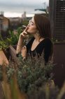 Side view of brunette posing sensually while smoking on balcony with cityscape on background. — Stock Photo